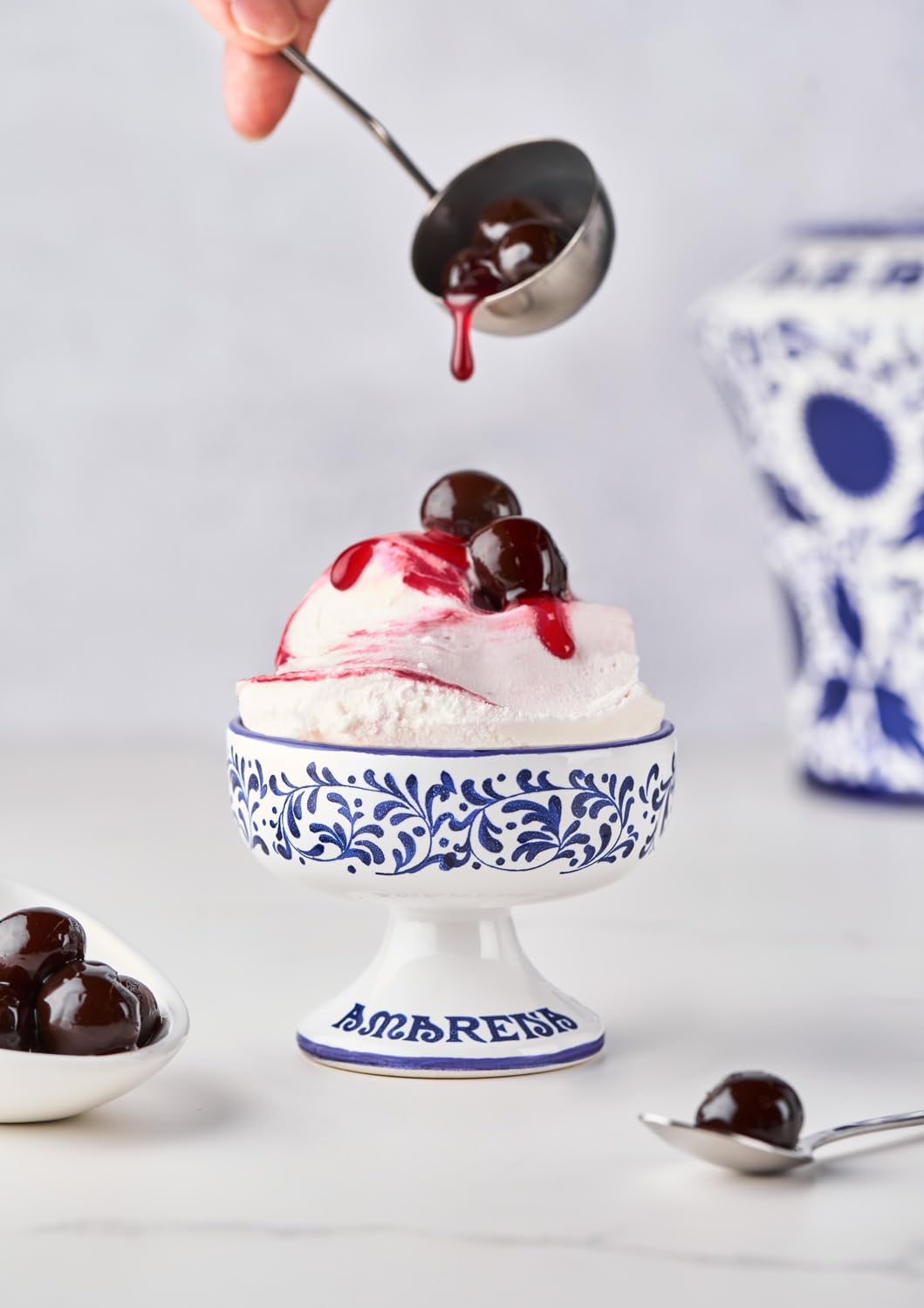 bowl of ice cream with amarena fabbri cherries scooped on top, bowl of cherries, and white and blue vases in background