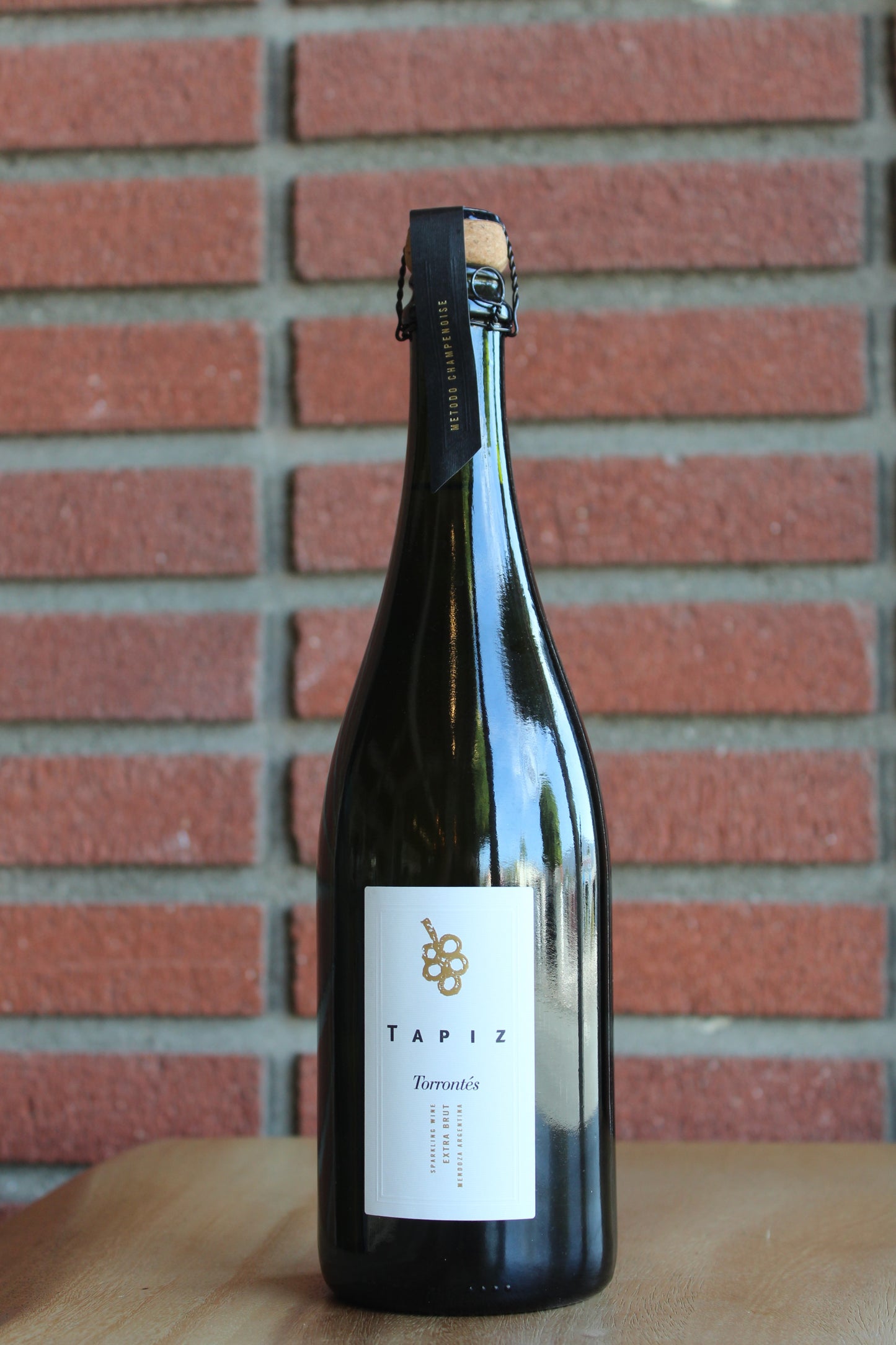 A dark glass champagne bottle of Tapiz sparkling Torrontes with a white label, standing on a wooden surface against a red brick wall background.