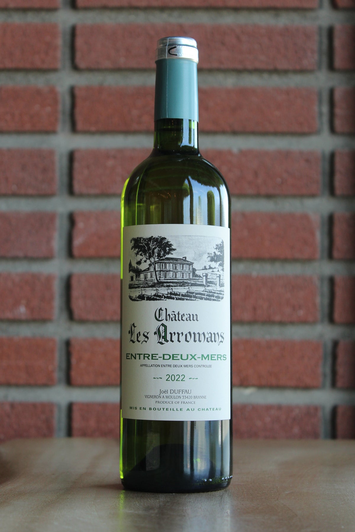 A bottle of Chateau Les Aromans Bordeaux Blanc - Entre Deux Mers white wine stands on a wooden surface against a red brick wall background.