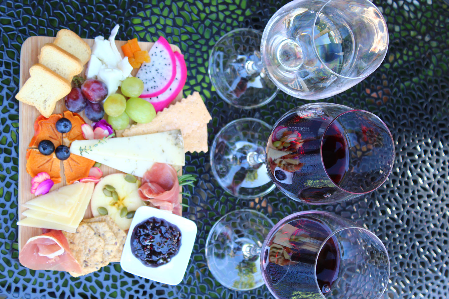 Platter of assorted cheeses, fruits, and meats with wine glasses on a textured surface