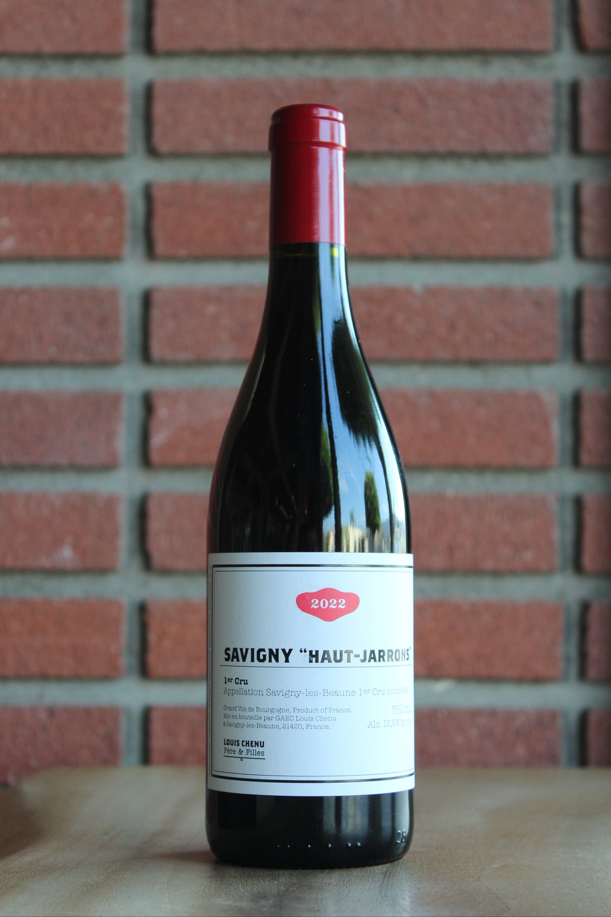 A bottle of Louis Chenu et Filles Savigny les Beaune "Haut-Jarrons" Premier Cru wine with a red foil top and white label, standing on a wooden surface against a red brick wall backgrou