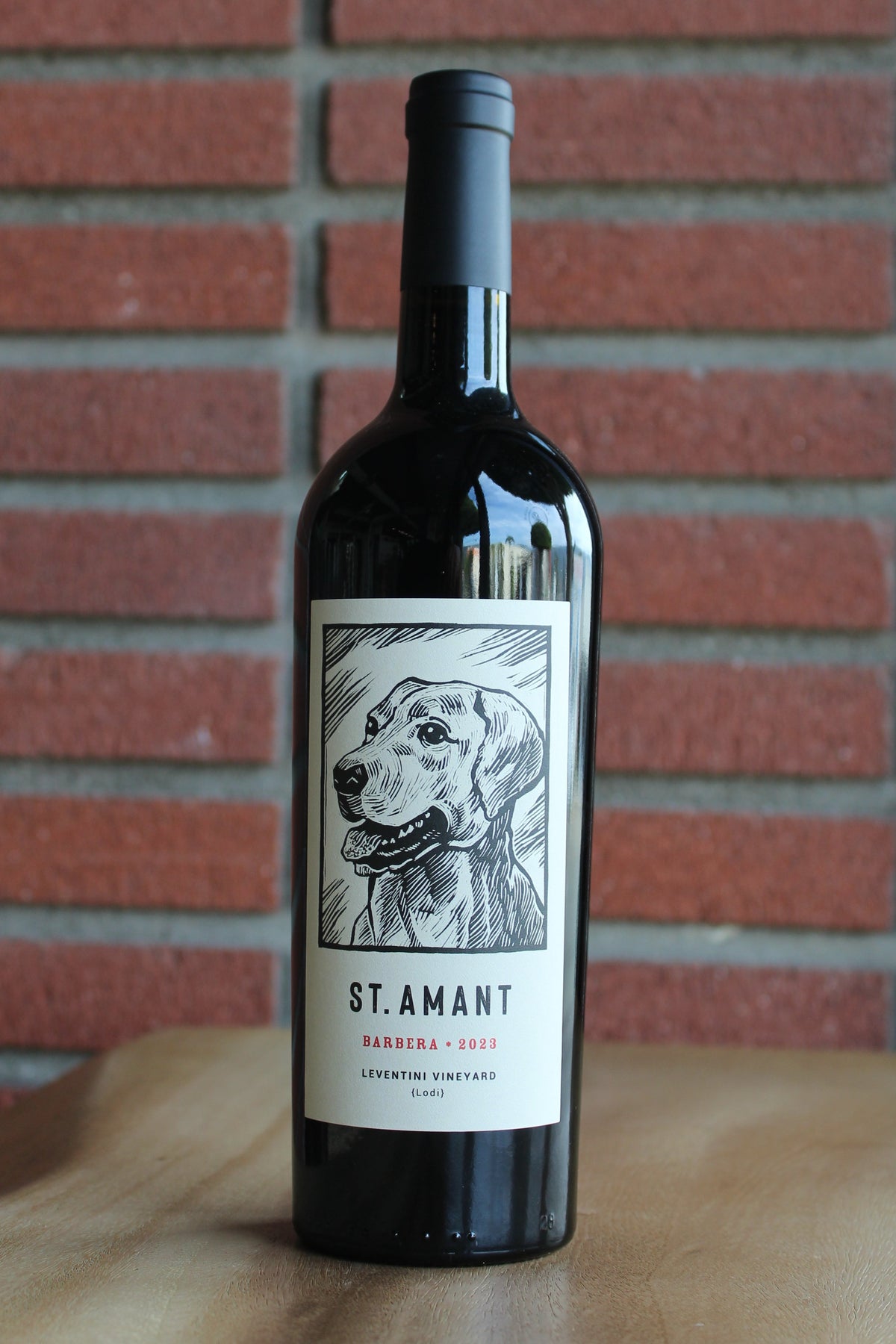 2023 St. Amant Barbera bottle from Leventini Vineyard in Lodi’s Jahant AVA on a wood surface with a red brick background