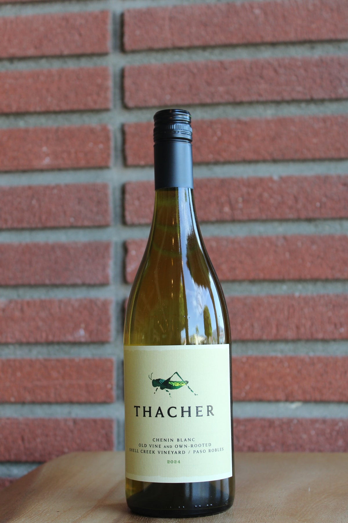 A bottle of Thacher Chinis Blanc dry wine with a cream label and black cap, standing on a wooden surface against a red brick wall background.
