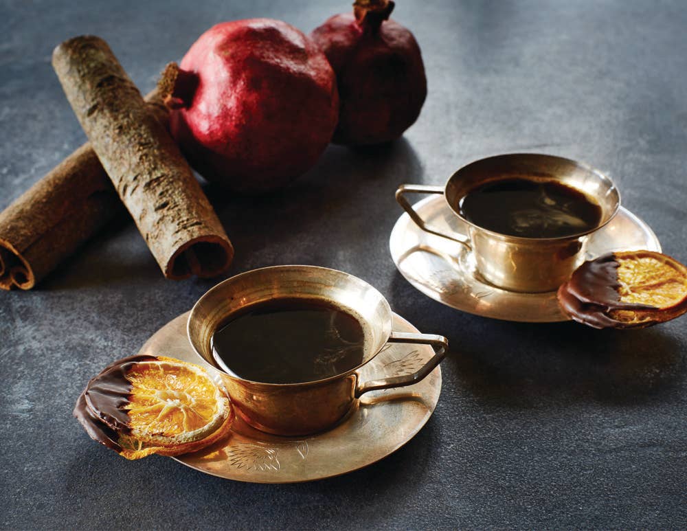 brass cups of a hot brown beverage with choclate dipped orang slices on the brass saucer with cinnamon sticks and pomegranates on a charcoal gray textured background