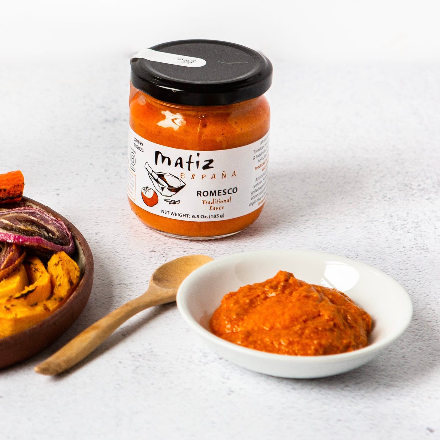 jar of matiz romesco, romesco in a bowl , some grilled vegetables on the side, all on a white background