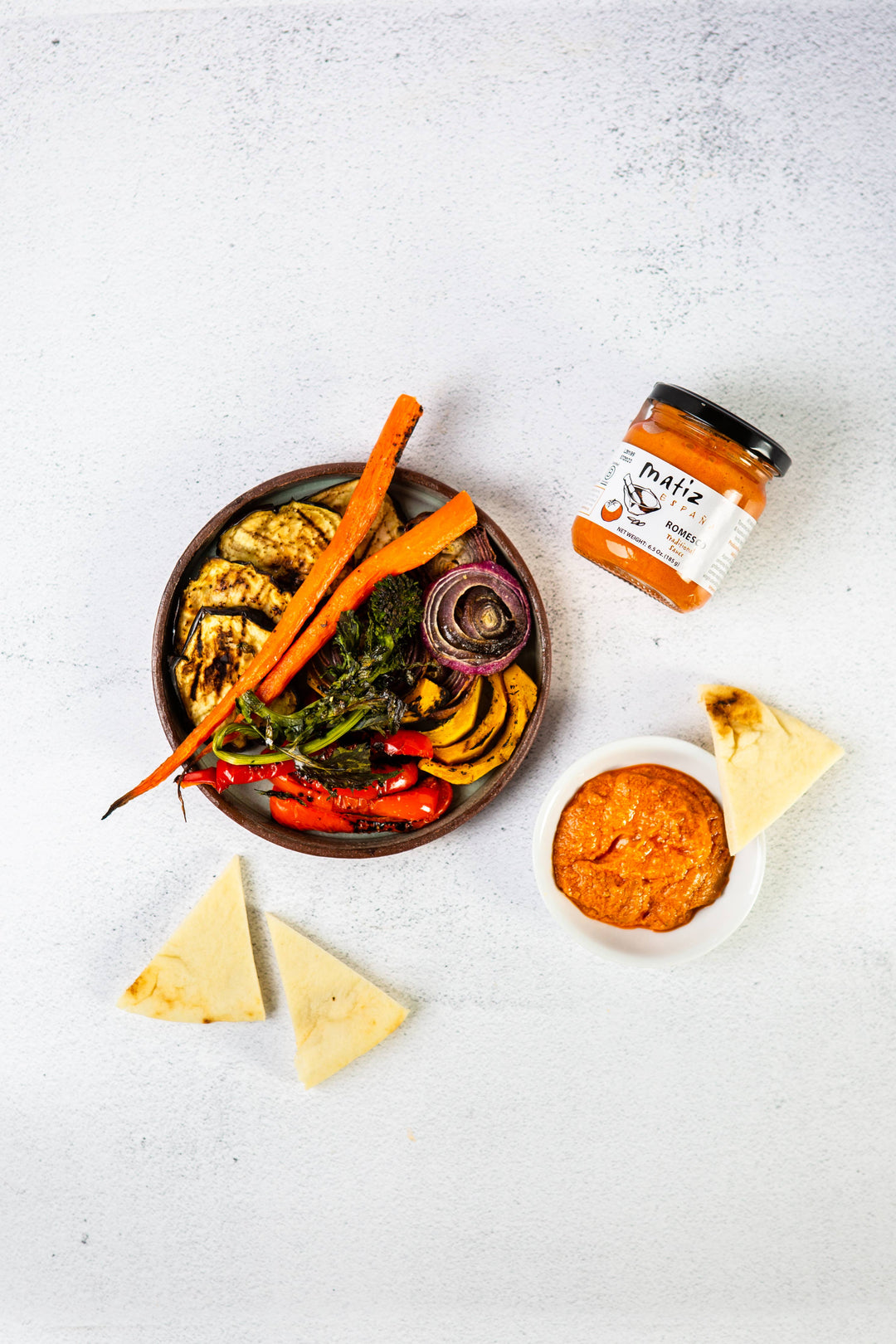 jar of matiz romesco, bowl of grilled vegetables, and pita slices on a white background
