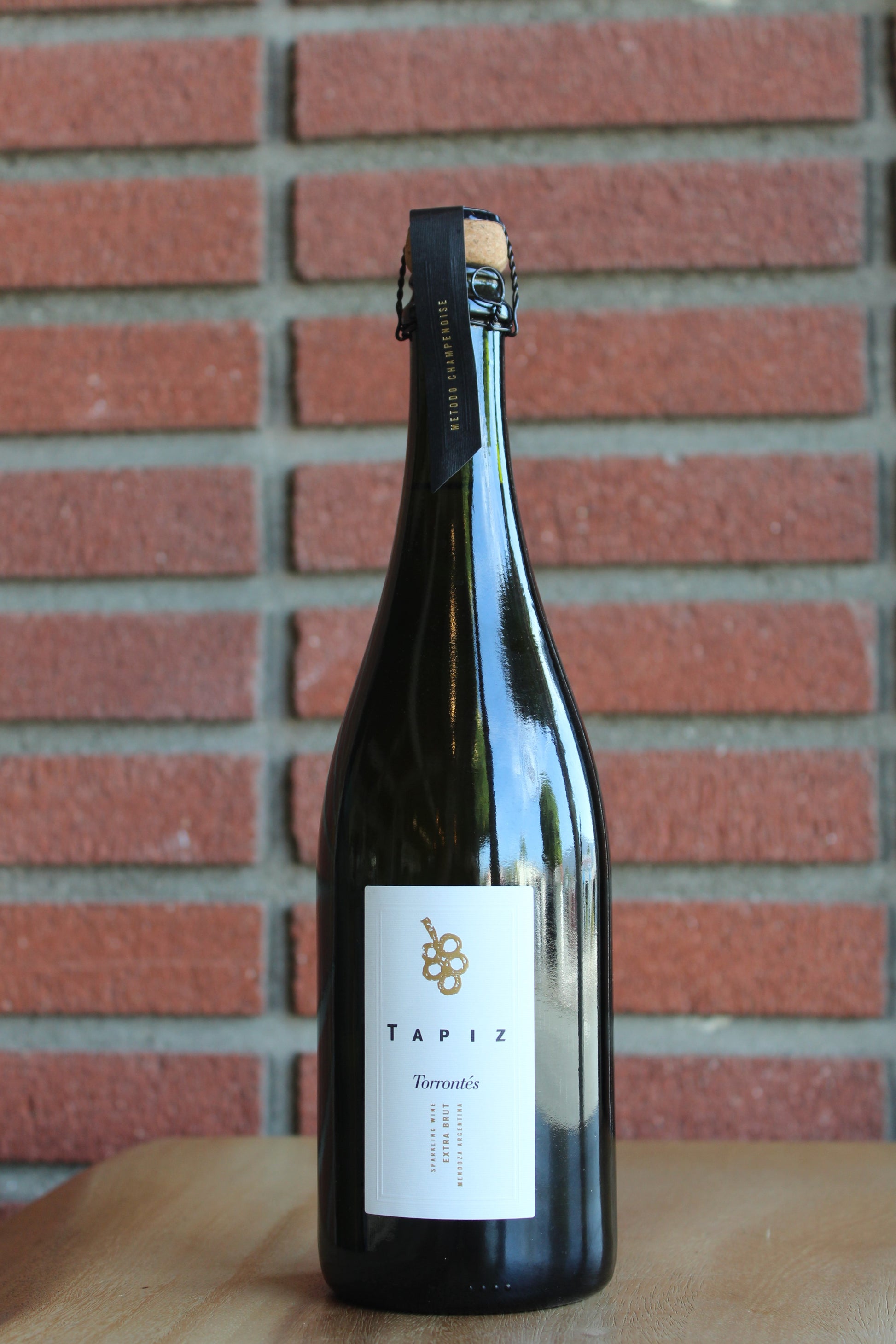 A dark glass champagne bottle of Tapiz  sparkling Torrontes with a white label, standing on a wooden surface against a red brick wall background.