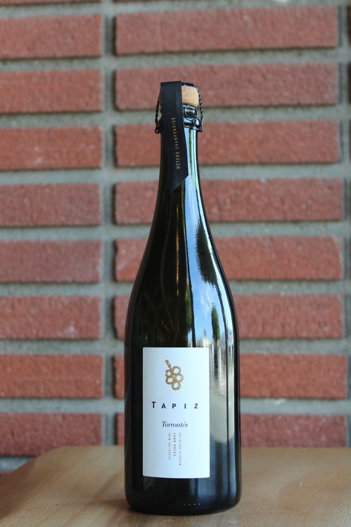 A dark glass champagne bottle of Tapiz  sparkling Torrontes with a white label, standing on a wooden surface against a red brick wall background.