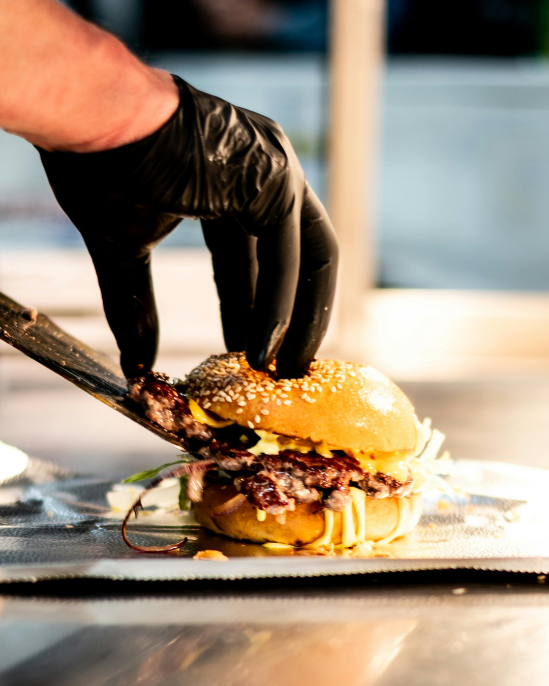 Hand with black glove holding a knife to cut a burger on a metal surface.