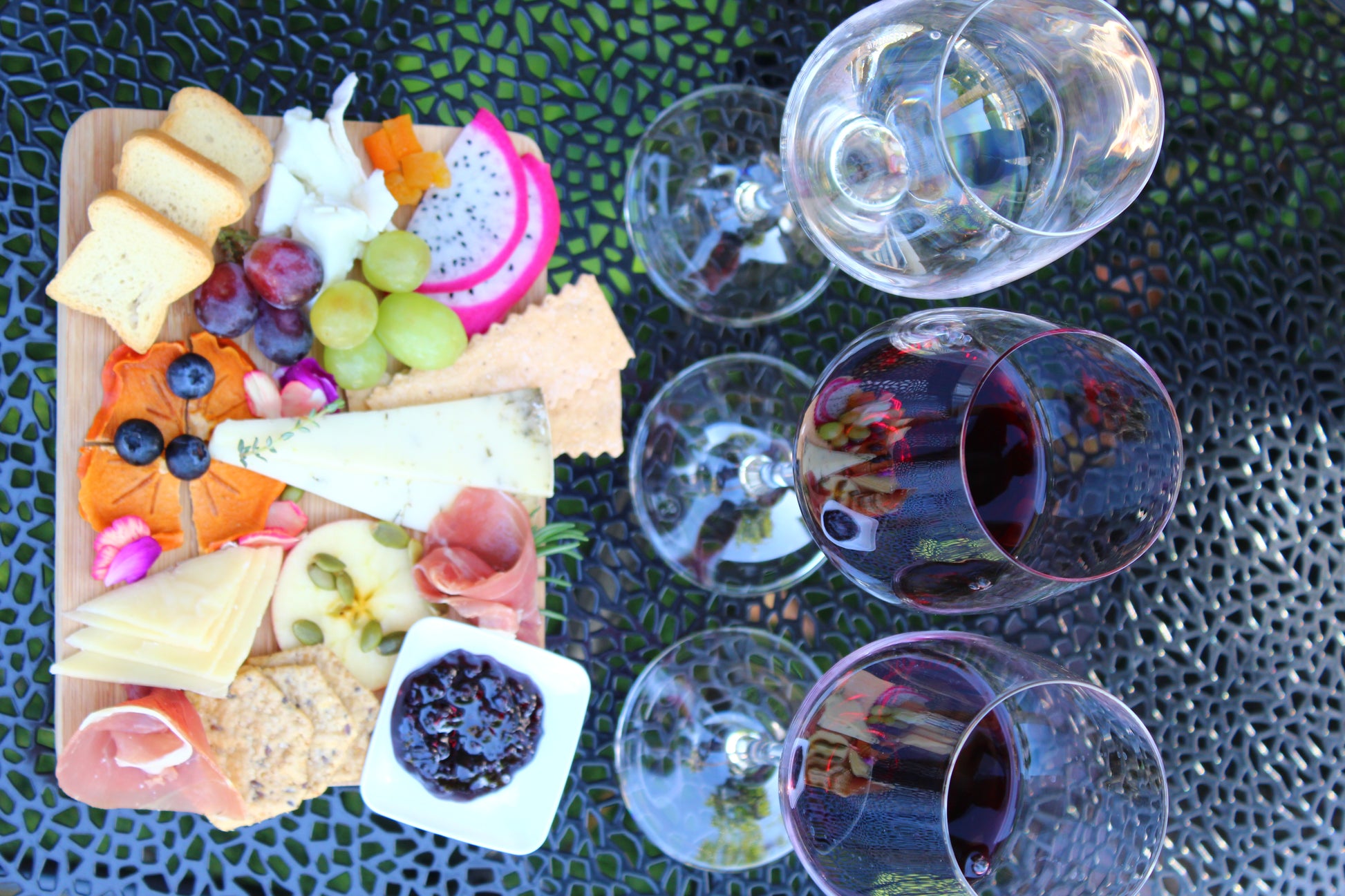 Platter of assorted cheeses, fruits, and meats with wine glasses on a textured surface