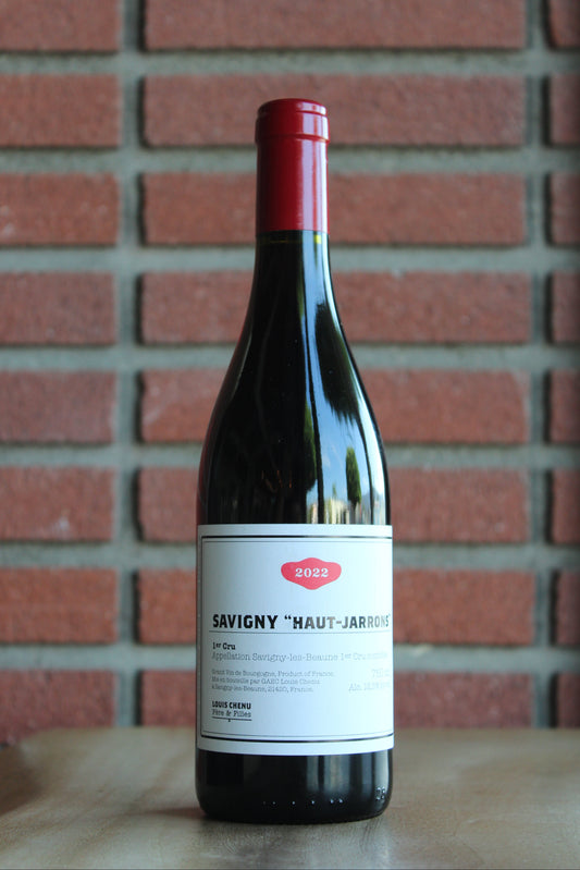 A bottle of Louis Chenu et Filles Savigny les Beaune "Haut-Jarrons" Premier Cru  wine with a red foil top and white label, standing on a wooden surface against a red brick wall backgrou