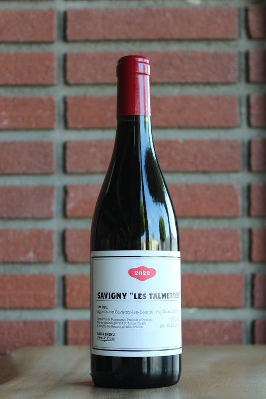 A bottle of Louis Chenu et Filles Savigny les Beaune " Les Talmettes" Premier Cru 2022 red wine with a red foil cap and white label, standing on a wooden surface against a red brick wall background.