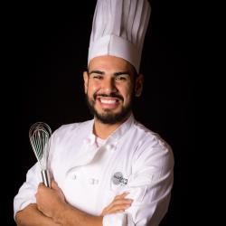 Chef Michael Mergill in a white uniform and tall hat holding a whisk against a black background