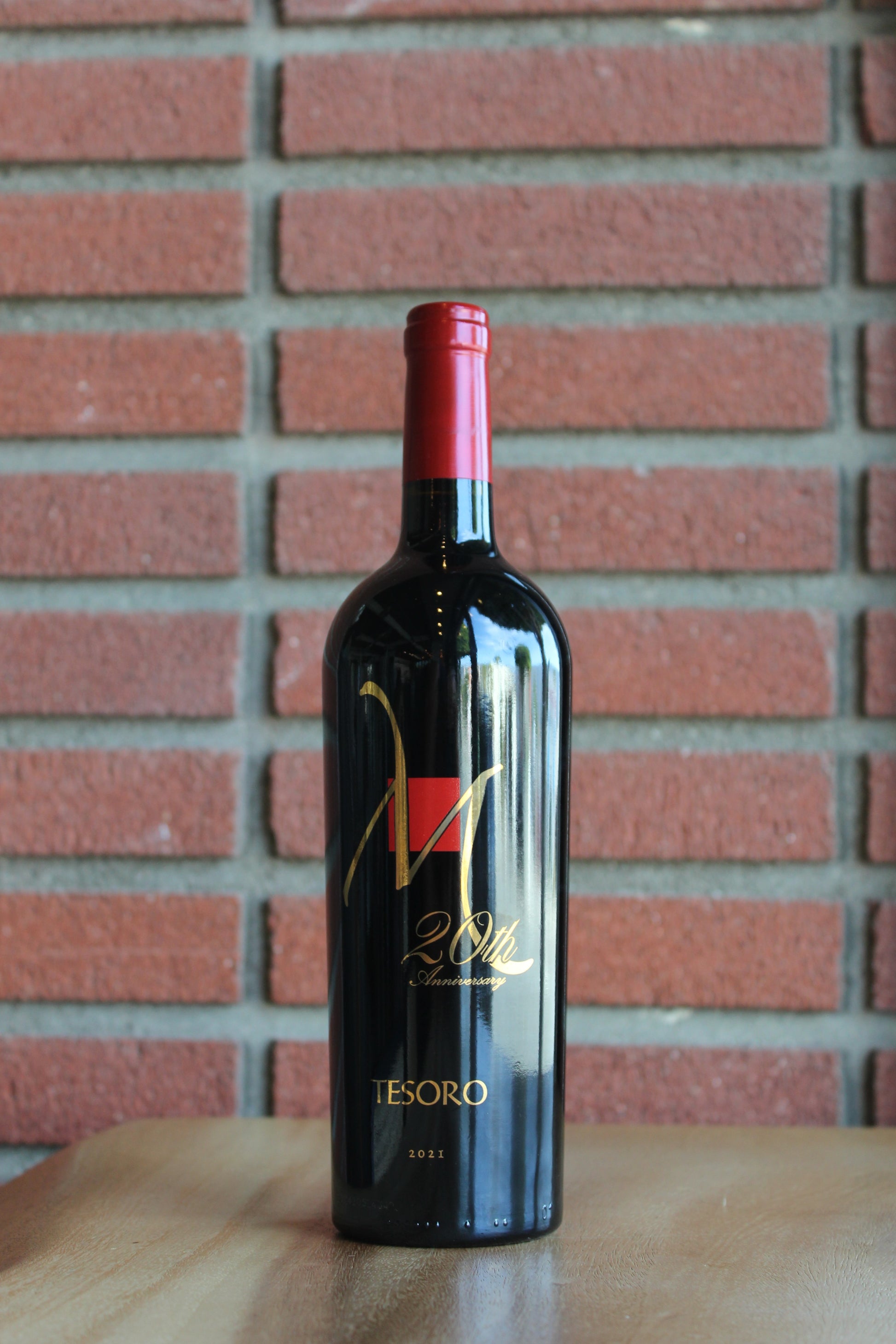 A bottle of Muscardini Cellars 20th Anniversary Tesoro red wine with a red foil top and gold lettering, standing on a wooden surface against a red brick wall background.