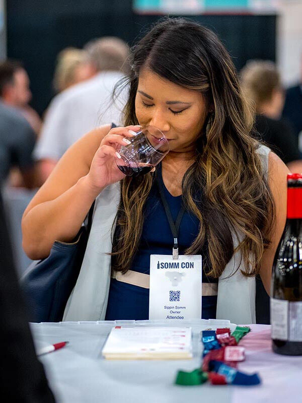 Ceritified Sommelier Kristina Tomlin drinking from a glass at a convention with a name tag and bottle of wine on a table.
