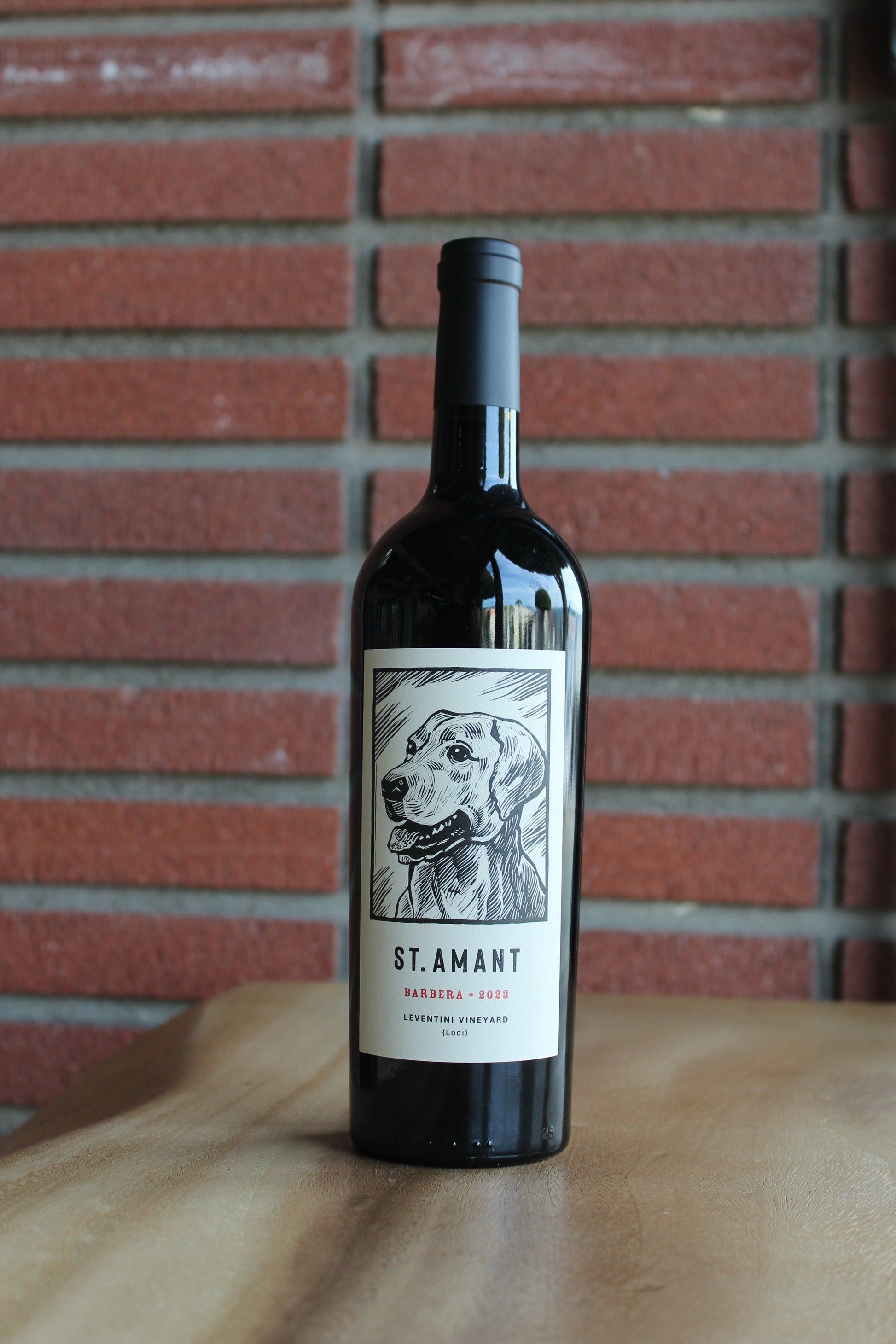 2023 St. Amant Barbera bottle from Leventini Vineyard in Lodi’s Jahant AVA  on a  wood surface with a red brick background