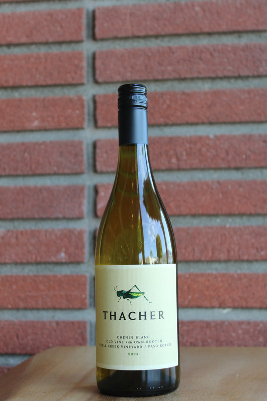 A bottle of Thacher Chinis Blanc dry wine with a cream label and black cap, standing on a wooden surface against a red brick wall background.