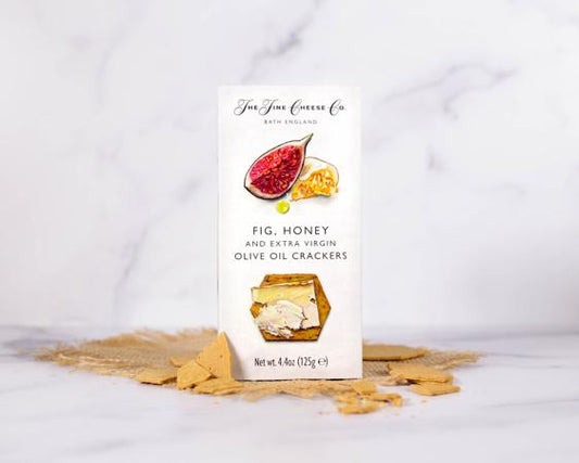 box of the fine cheese co fig, honey, and olive oil on a white marble background with crackers scattered